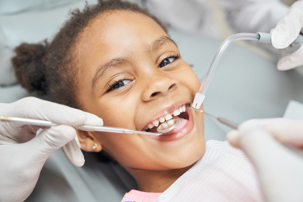 Why Kids Get Cavities Even When Parents Are Doing Everything Right, Charming little afro american girl sitting in dental chair, smiling and looking at camera during medical treatment at modern clinic. Concept of health care ad pediatrics